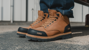 A person wearing blue jeans and tan Xero Shoes lace-up boots with black toe caps stands on a concrete surface outdoors, showcasing comfortable work boots perfect for any task.